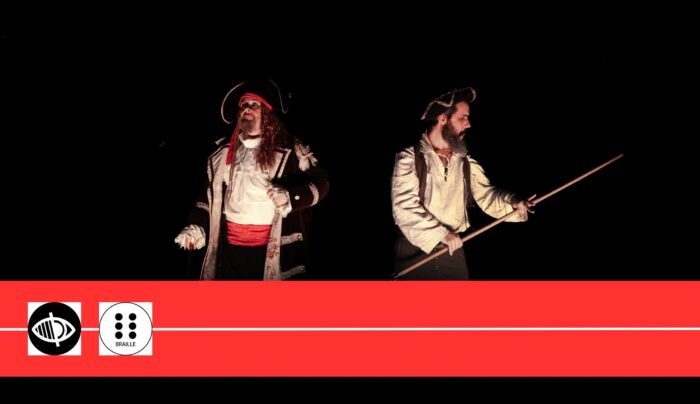 Dos actors caracteritzats com a pirates interpreten una escena sobre un escenari fosc. Un d’ells sosté una llança mentre l’altre gesticula, tots dos il·luminats per focus puntuals.