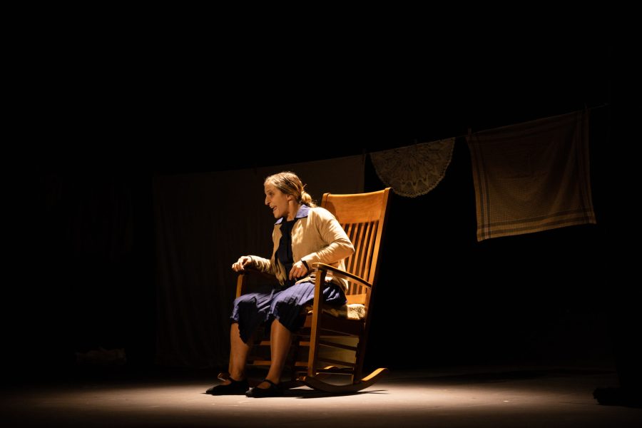 Una actriu asseguda en una cadira de balancí interpreta una escena sobre un escenari gairebé buit. Al fons, un estenedor amb llençols i peces de roba reforça l’ambient domèstic de l’escenografia.