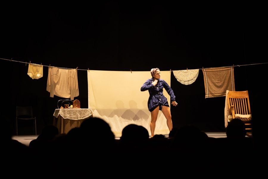 Una actriu interpreta una escena al centre de l’escenari davant d’una escenografia que recrea un espai domèstic. Un estenedor amb roba, una taula amb estovalles i una cadira de balancí emmarquen l’acció.