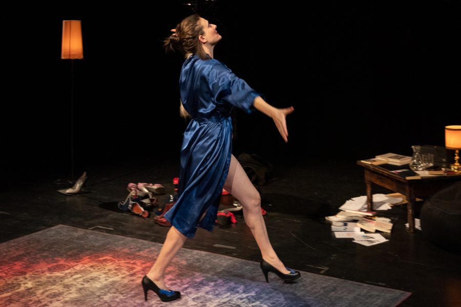 Una dona travessa l’escenari amb una bata blava i sabates de taló. Amb els braços oberts i el cos en moviment, sembla expressar una emoció intensa en un espai que recrea un camerino teatral.
