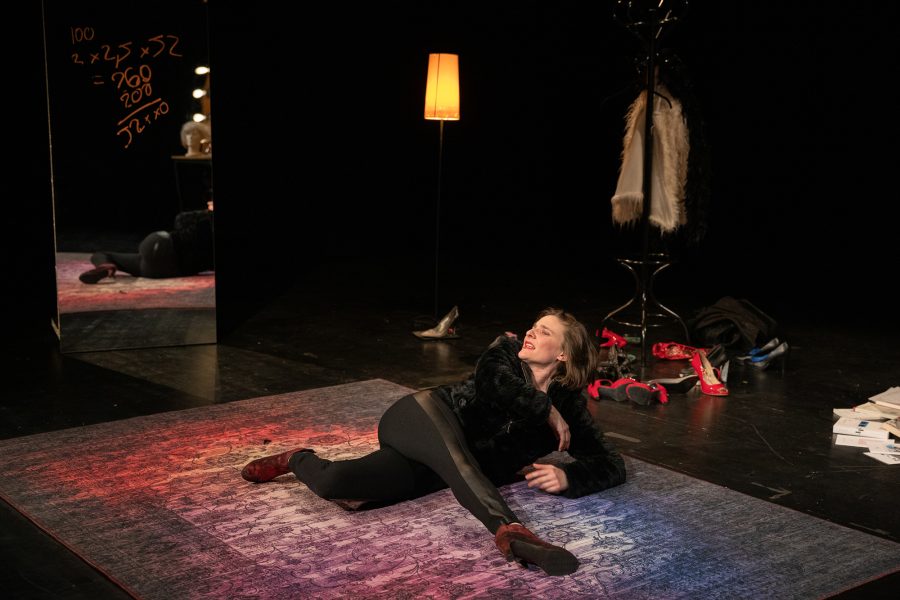 na dona està estirada sobre una catifa al centre de l’escenari. Al seu voltant hi ha sabates, peces de roba i elements de camerino que suggereixen un moment de vulnerabilitat o esgotament.