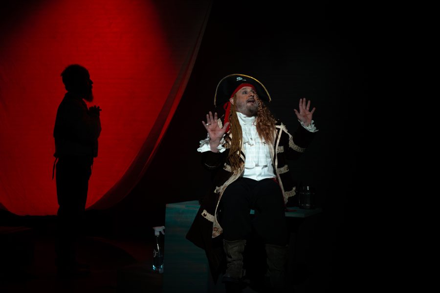 Un actor caracteritzat com un pirata, amb tricorni i cabells llargs, interpreta una escena sobre un escenari fosc. Al fons, la silueta d’una altra persona es retalla davant d’una gran llum vermella.