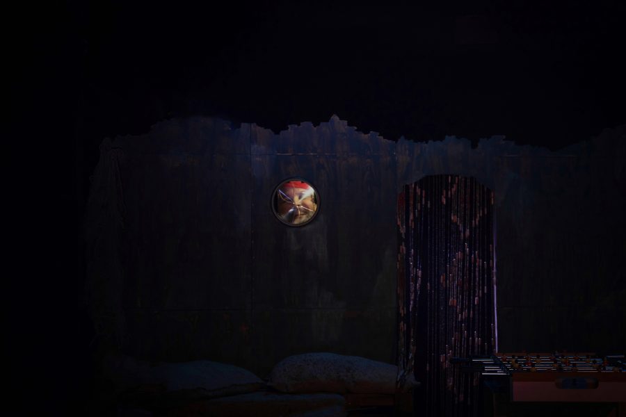 Escenografia d’un espectacle amb una estructura fosca que recorda una cabina o un refugi. Una petita finestra circular deixa entreveure una figura a l’interior, mentre una cortina de tires brillants marca una entrada lateral