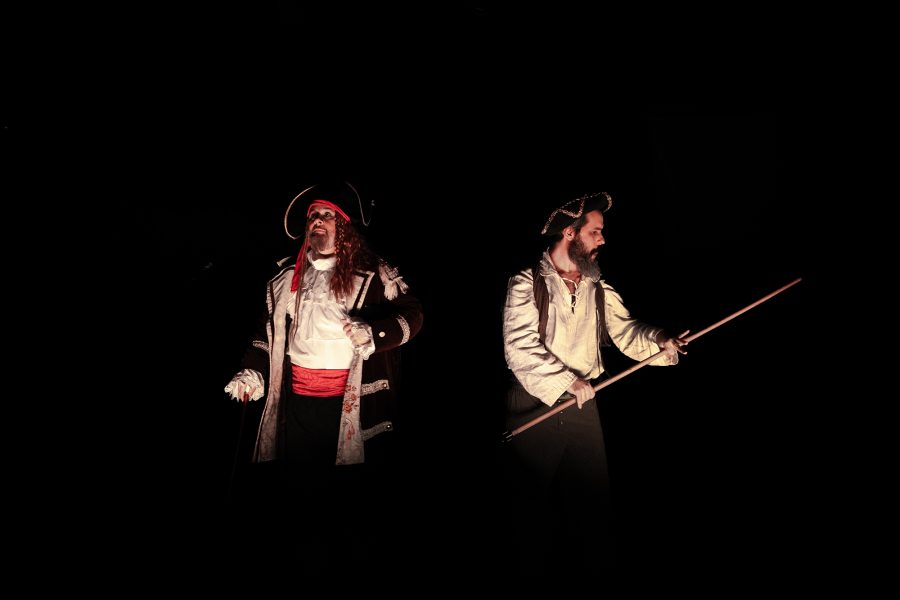 Dos actors caracteritzats com a pirates interpreten una escena sobre un escenari fosc. Un d’ells sosté una llança mentre l’altre gesticula, tots dos il·luminats per focus puntuals.