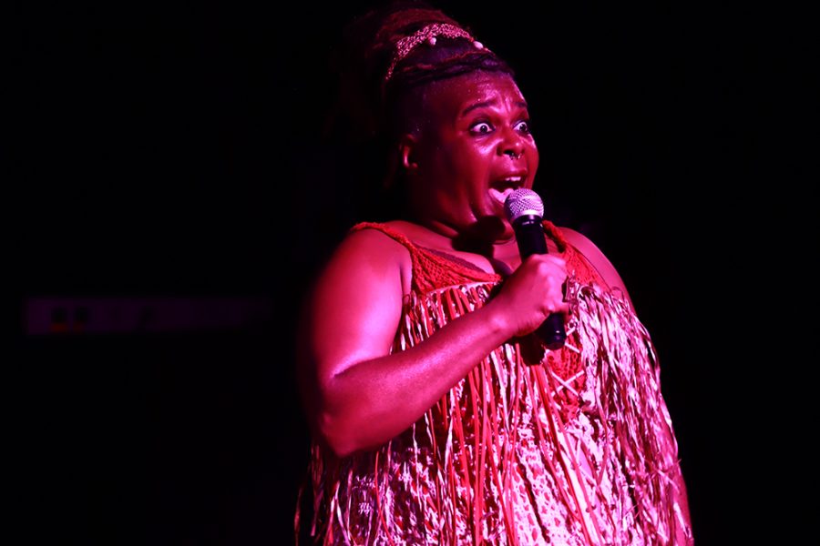 Una dona afrodescendent canta o parla davant d’un micròfon sobre un escenari fosc. Vestida amb un vestit de serrells brillants, mira el públic amb una expressió intensa i enèrgica.