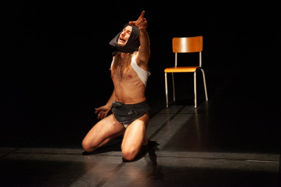 Un actor agenollat al centre d’un escenari fosc assenyala cap amunt amb un braç estès. Porta el cap embolcallat amb una peça de roba i, al seu darrere, una cadira buida queda il·luminada per un focus.