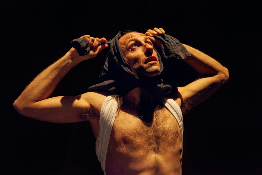 Un actor amb el tors descobert sosté una peça de roba fosca sobre el cap mentre mira cap amunt. La il·luminació teatral destaca la tensió del gest i l’expressió intensa del seu rostre.