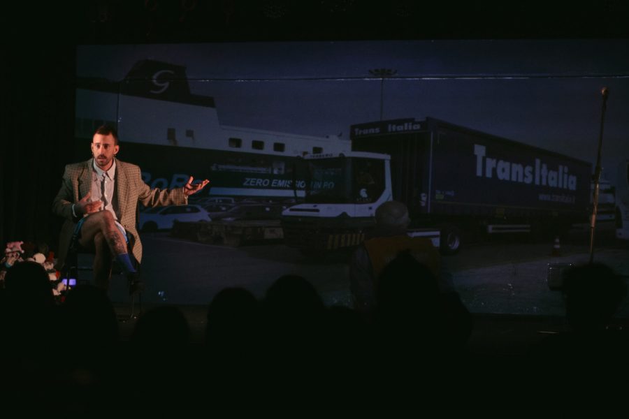 Una persona asseguda en una cadira parla al públic sobre un escenari mentre al fons es projecta la imatge d’un camió de transport. L’escena combina narració en directe i projecció audiovisual.