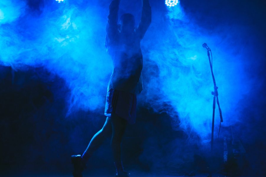 Una persona apareix a contrallum sobre un escenari envoltat de fum i il·luminat amb llum blava. Amb els braços alçats sobre el cap, adopta una postura expressiva davant d’un micròfon.
