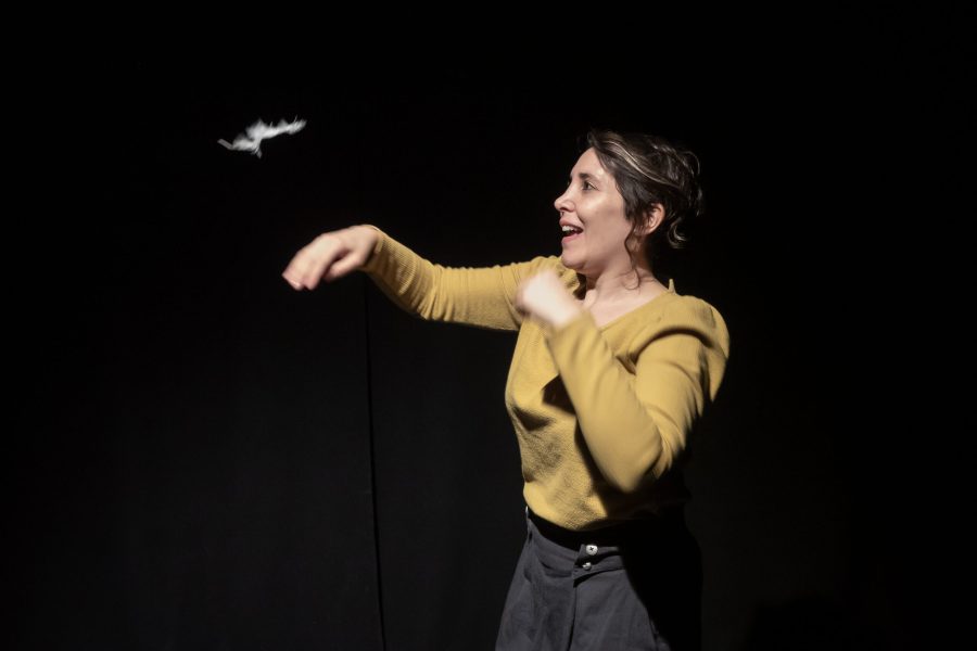Una actriu sobre un escenari fosc gesticula amb els braços mentre observa una ploma blanca suspesa a l’aire. La seva expressió transmet sorpresa i curiositat.