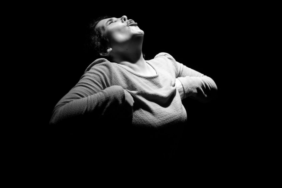 Una actriu il·luminada per un focus emergeix de la foscor amb el cap inclinat cap enrere i els braços flexionats. La imatge en blanc i negre transmet una gran intensitat física i emocional.
