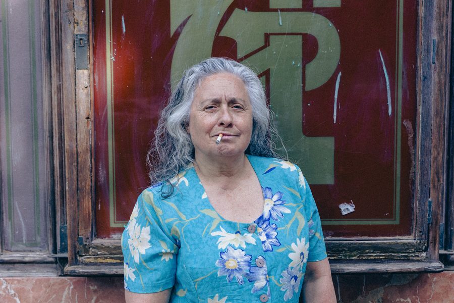 Una dona gran amb els cabells grisos fuma una cigarreta davant de l’aparador d’un estanc. Mira a càmera amb una expressió segura.