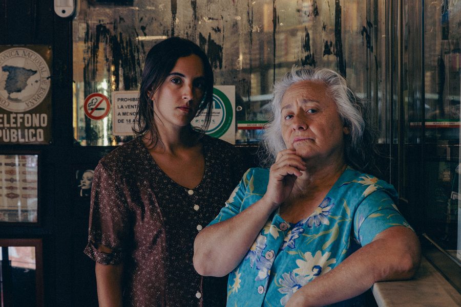 Dues dones es troben a l’interior d’un estanc tradicional. Una dona gran amb cabells blancs recolza el braç al taulell mentre una dona més jove l’acompanya al seu costat. Ambdues miren a càmera amb expressió seriosa.