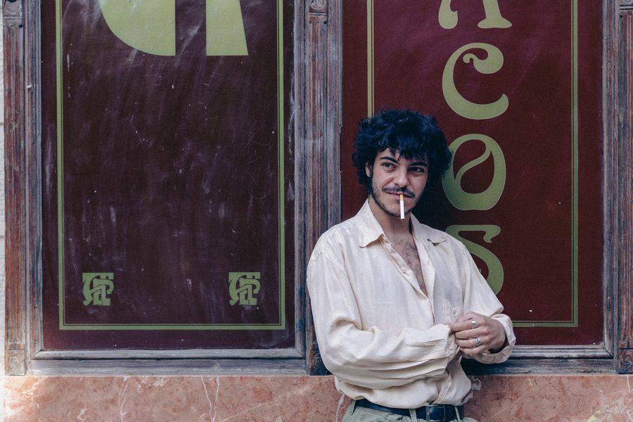 Un jove amb una cigarreta als llavis recolza l’esquena contra l’aparador d’un estanc. Mira cap a un costat amb una expressió relaxada mentre es corda la camisa.