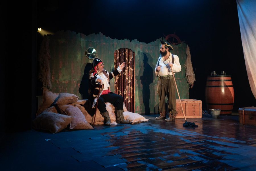 Dos actors caracteritzats com a pirates conversen sobre un escenari que recrea un moll o refugi costaner. L’espai inclou sacs, caixes, un barril i una construcció de fusta envellida.