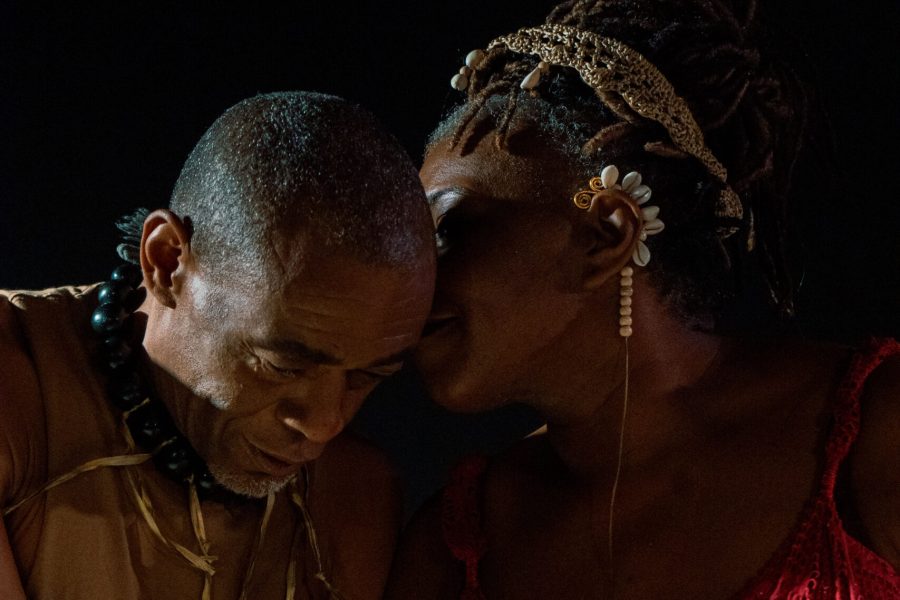 Dues persones afrodescendents comparteixen un moment d’intimitat sobre l’escenari. Una d’elles s’acosta a l’orella de l’altra com si li xiuxiuegés alguna cosa mentre mantenen els caps junts.