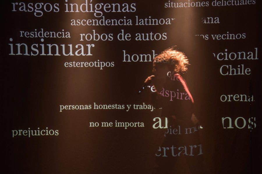 Una actriu apareix parcialment il·luminada sobre l’escenari mentre sosté un micròfon. Sobre el seu cos i el fons es projecten paraules i expressions en castellà relacionades amb prejudicis, estereotips i migració, creant una superposició visual entre el text i la figura.