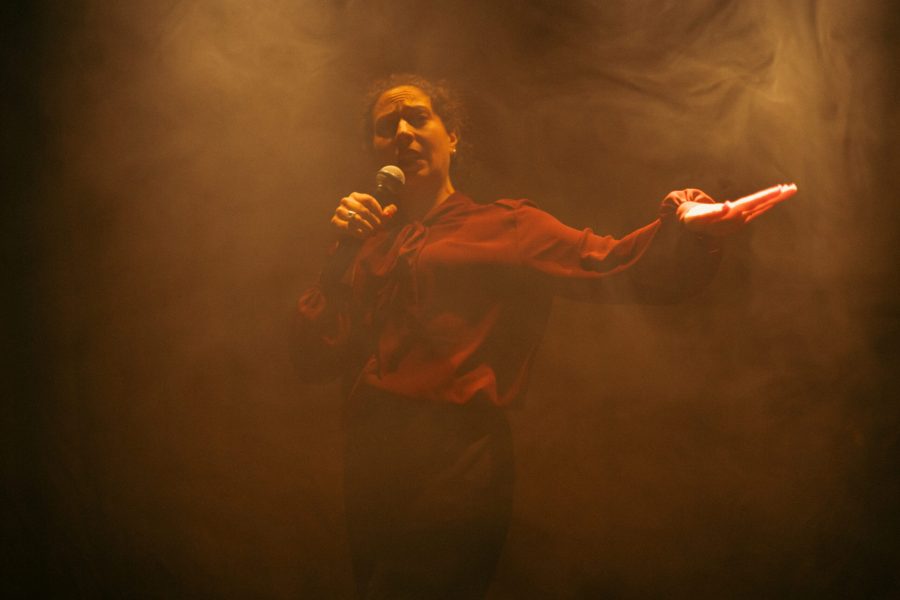 Una actriu sosté un micròfon amb una mà mentre estén l’altra davant seu sobre un escenari envoltat de fum. La il·luminació càlida crea una atmosfera intensa i difumina el fons de la imatge.