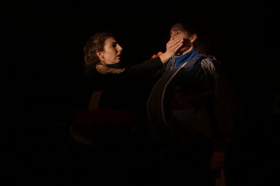 Una actriu tapa la boca d’una altra dona sobre un escenari fosc. La il·luminació focalitza el gest i crea una escena de tensió i confrontació.