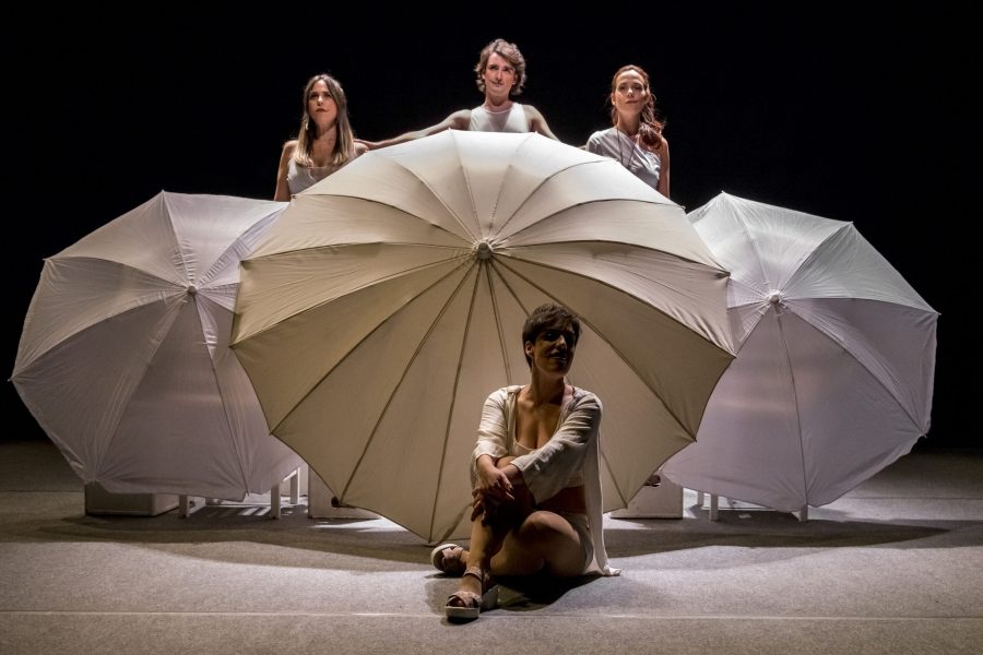 Tres actrius i un actor ocupen diferents nivells de l’escenari envoltades de grans paraigües blancs oberts. Una d’elles seu al davant mentre les altres tres es mantenen dretes al darrere, creant una composició simètrica i serena.