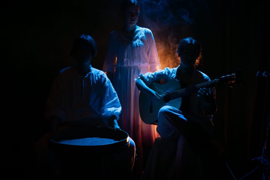 Tres intèrprets vestits de blanc ocupen l’escenari sota una il·luminació blava i ataronjada. Una persona toca la guitarra, una altra seu davant d’un instrument de percussió i una tercera roman dreta al centre envoltada de fum.