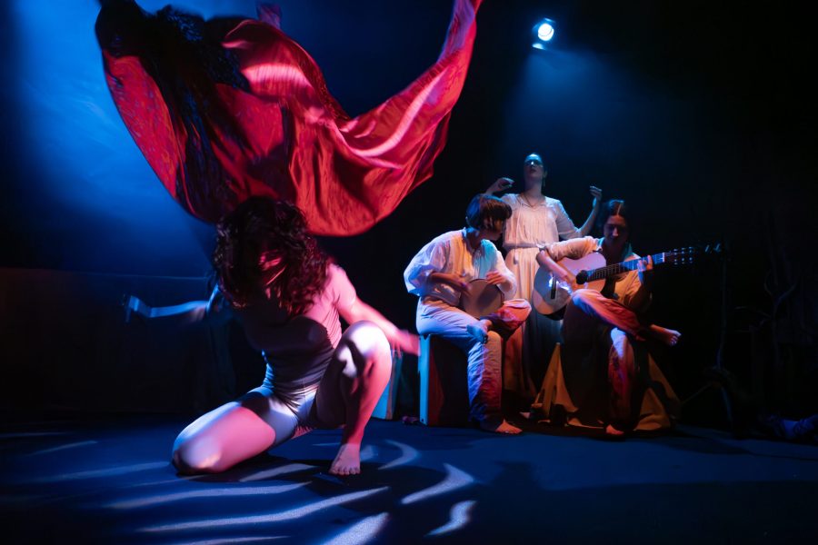 Una actriu agenollada al primer terme es mou sota una gran tela vermella que vola per l’aire. Al fons, tres intèrprets vestits de blanc acompanyen l’escena amb música i cant sota una il·luminació blava i vermella.