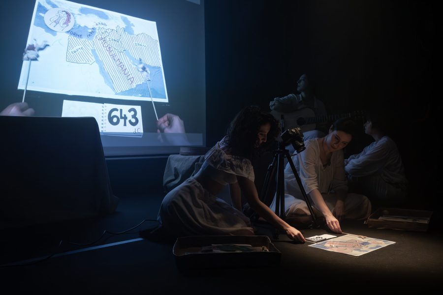 Diversos intèrprets treballen al voltant de documents estesos a terra mentre una projecció mostra un mapa i material d’arxiu al fons de l’escenari. La imatge combina investigació, narració visual i música en directe.