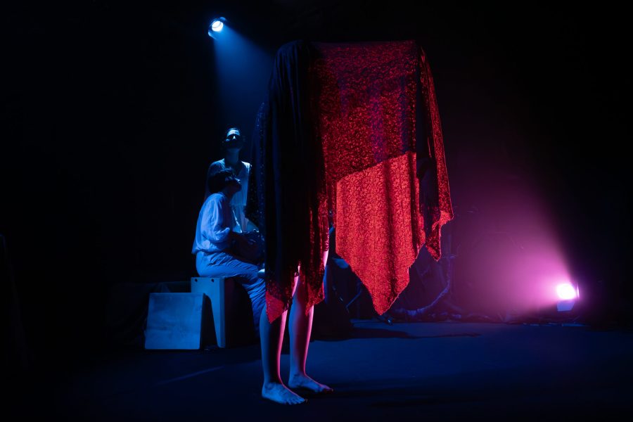 Una figura dempeus al centre de l’escenari queda completament coberta per una tela vermella translúcida que cau fins als peus. Al fons, altres intèrprets romanen en penombra sota una il·luminació blava i magenta que crea una atmosfera misteriosa.