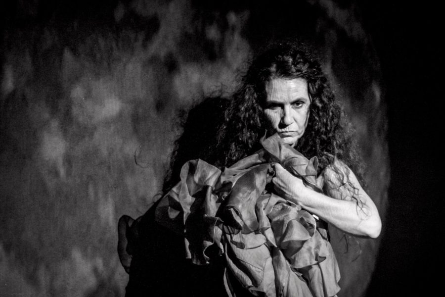 Una actriu amb els cabells llargs i arrissats sosté una gran peça de tela arrugada contra el cos. En una imatge en blanc i negre, mira directament a càmera amb una expressió intensa davant d’un fons que evoca la superfície de la lluna.